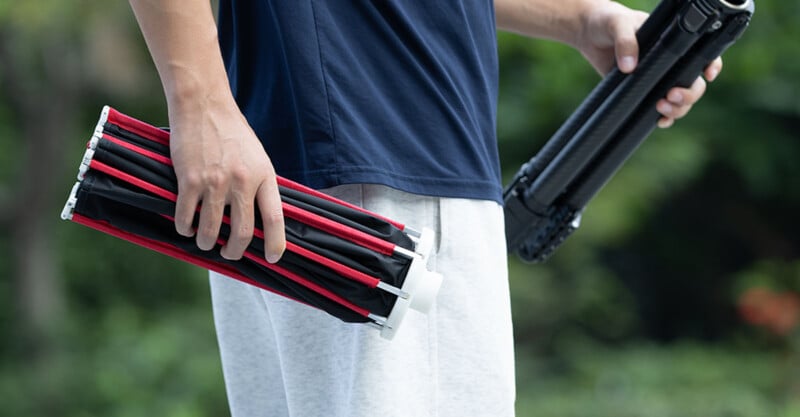A person in a blue t-shirt and grey sweatpants holds a folded black and red photography softbox in one hand and a tripod in the other.