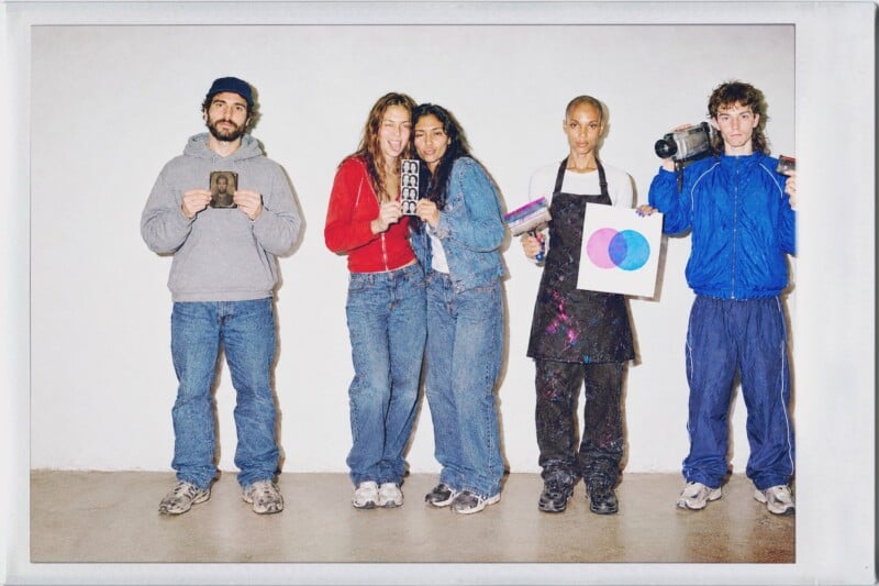 Four people stand in a line against a white wall, holding various objects including a small portrait, a photo strip, an art print, and a video camera.