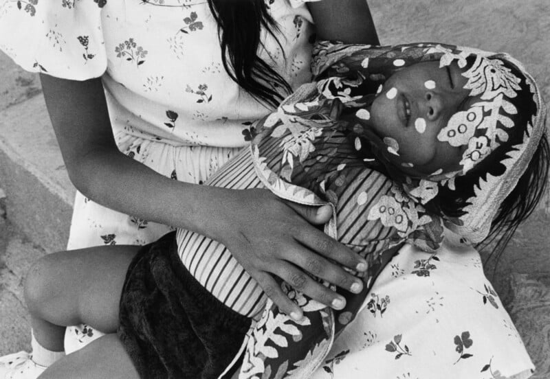 A person in a floral dress holds a sleeping child whose face is partially covered by a patterned headscarf.