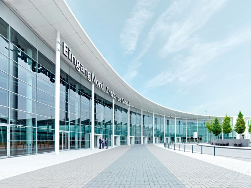 A modern, curved glass building with the sign "Eingang Nord Entrance North" features a wide paved walkway leading to its entrance, where three people stand.