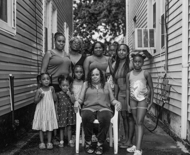 A group of ten Black women and girls of various ages pose for a portrait in a narrow alleyway between two houses.