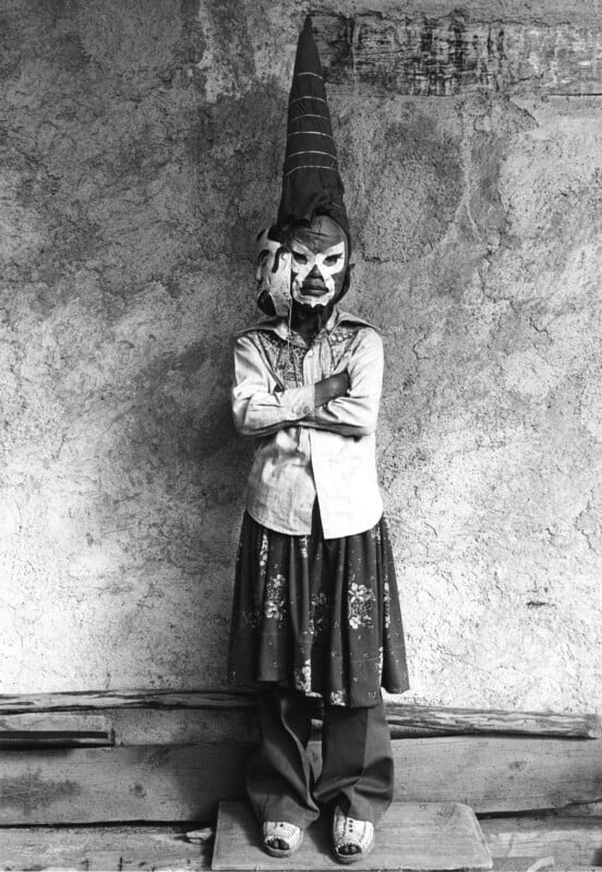 A person stands with arms crossed against a textured wall, wearing a tall, conical headpiece, a mask with white markings, a button-down shirt, a floral skirt over trousers, and sandals.