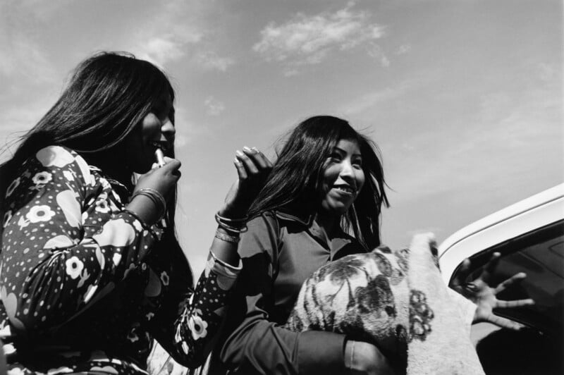 Two young women with long dark hair stand outdoors, one applying lipstick while the other smiles and holds a patterned fabric.