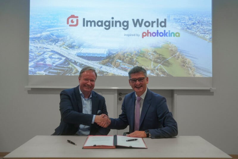 Two men in suits shake hands over a signed document on a table in front of a projection screen displaying the text "Imaging World inspired by photokina.