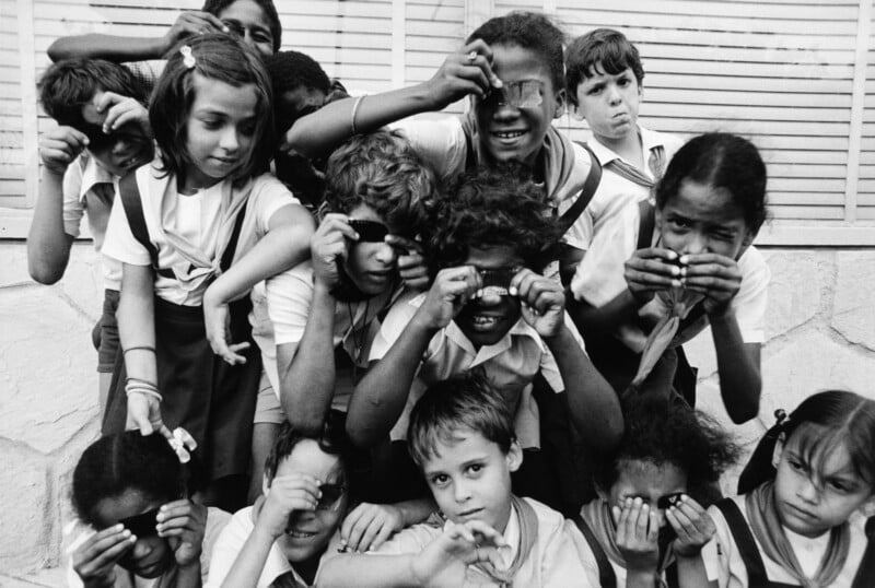 A group of children in school uniforms look through strips of film negatives held up to their eyes.