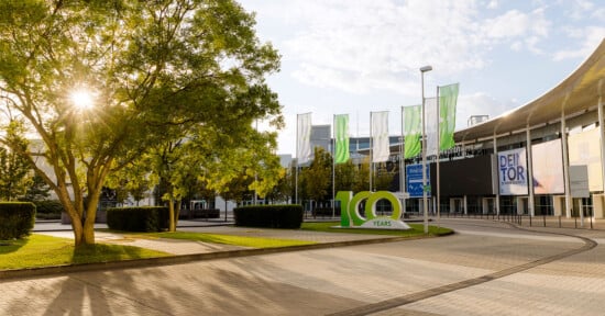 A large "100 YEARS" sign stands on a paved plaza in front of a modern building with green flags and banners under a sunny sky.