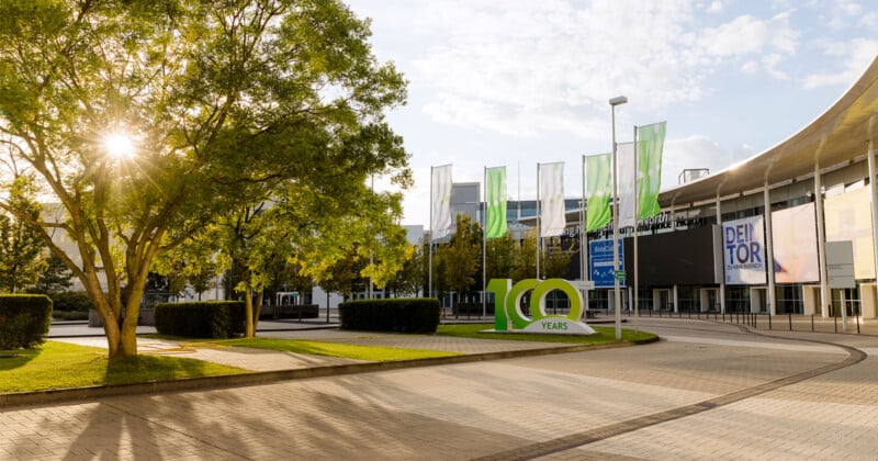 A large "100 YEARS" sign stands on a paved plaza in front of a modern building with green flags and banners under a sunny sky.