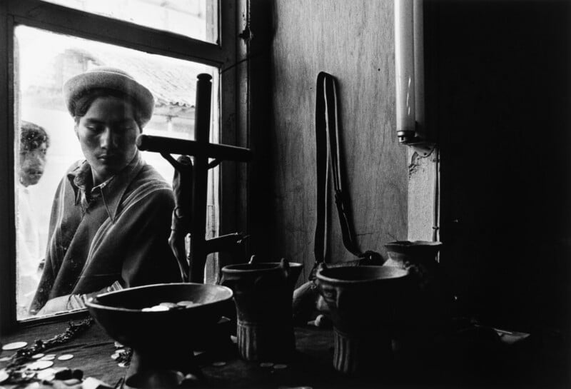 A young man in a hat looks through a window at a collection of ceramic bowls and a crucifix on a wooden surface.