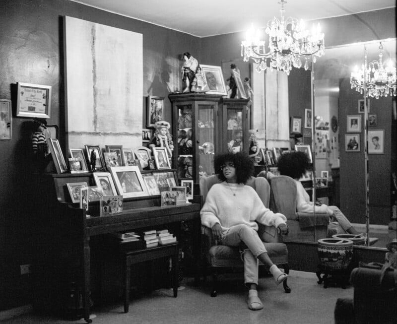 A person with an afro sits in an armchair in a room filled with framed photos, a piano, a glass display cabinet, and a large mirror reflecting the scene.
