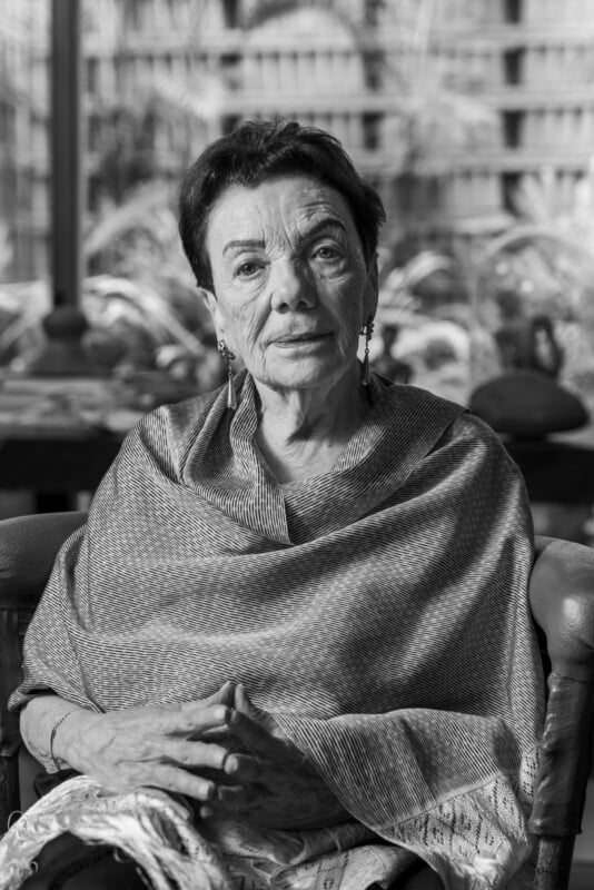 A portrait of an elderly woman with short dark hair, wearing a striped shawl and dangling earrings, sitting with her hands clasped.