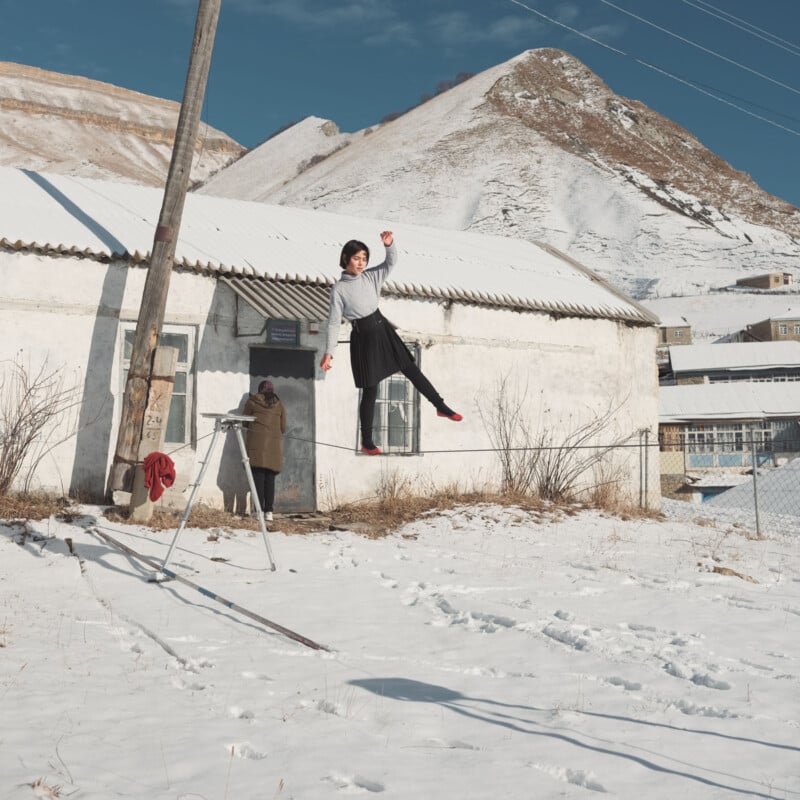 A person balances on a tightrope in front of a white building, with a snow-covered mountain landscape in the background.