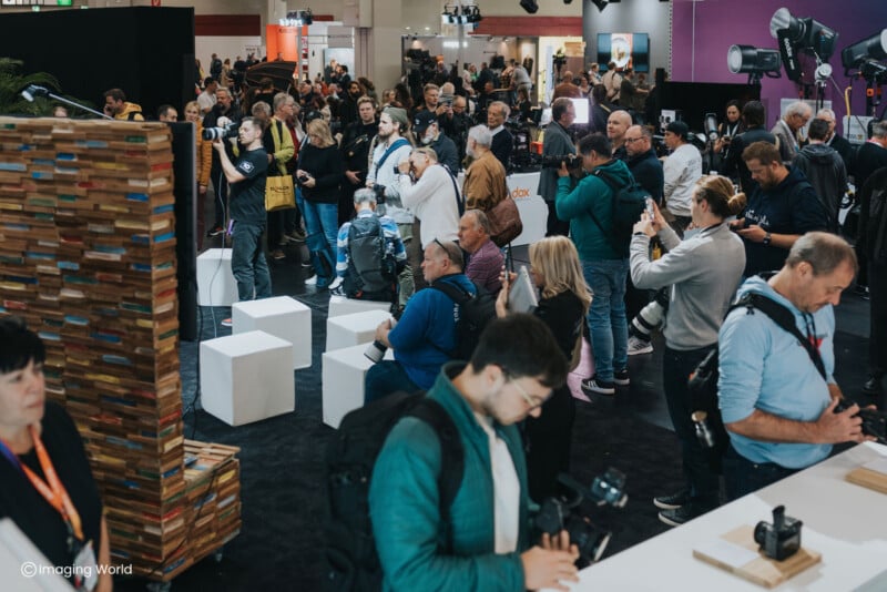 A crowded trade show floor features many people holding cameras and looking at displays, with white cube seating scattered throughout.
