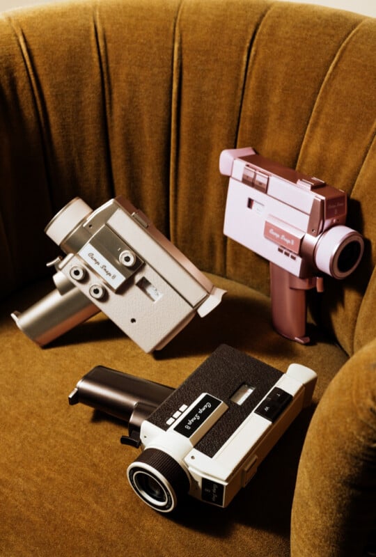 Three vintage-style video cameras in white, pink, and black sit on a textured, mustard-yellow velvet chair.