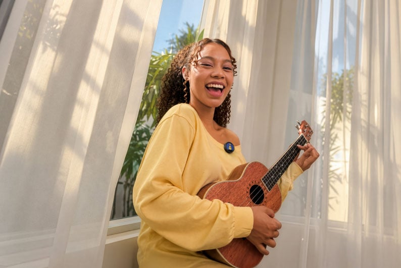 A smiling woman with curly hair wears a yellow long-sleeved shirt and plays a wooden ukulele while standing near a window with sheer curtains.
