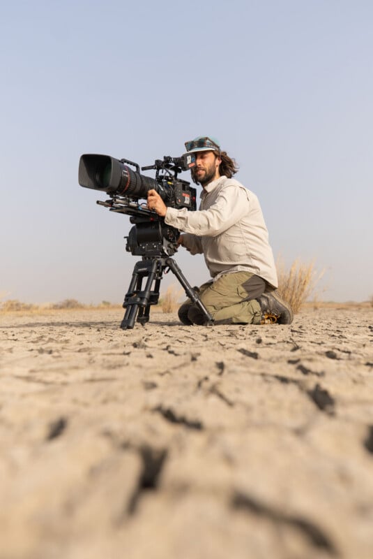 A man with long hair and a cap kneels on cracked, dry earth while operating a professional video camera mounted on a tripod.