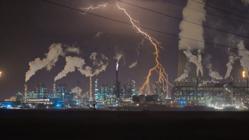 A lightning bolt strikes near an industrial plant at night, with smoke rising from multiple chimneys and cooling towers.