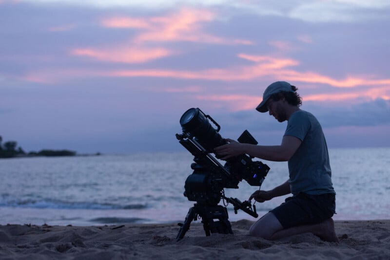 A person wearing a cap and t-shirt kneels on a sandy beach at sunset, adjusting a professional video camera mounted on a tripod.