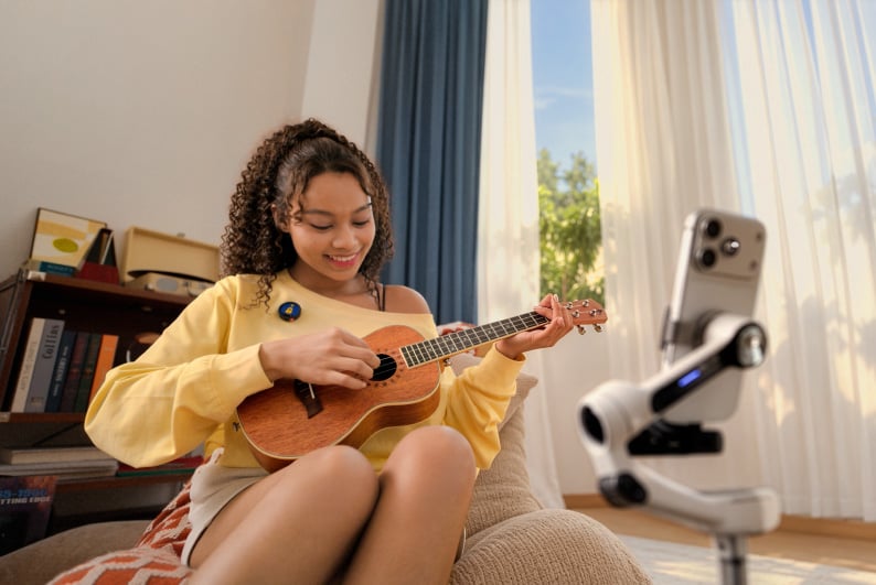 A young woman with curly hair sits on a beanbag chair, smiling while playing a ukulele in front of a smartphone mounted on a gimbal.