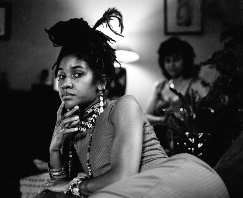 A young woman with dreadlocks styled upward with a feather poses while reclining, wearing a cowrie shell necklace and bracelets, with a blurred figure in the background.