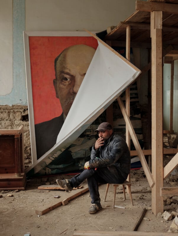 A man in work clothes sits on a small stool smoking a cigarette in a dusty, unfinished room next to a large, partially covered portrait of Vladimir Lenin.