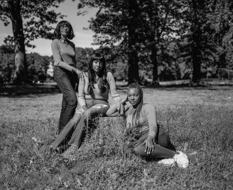 Three Black women of different generations pose together in a grassy park, with one standing, one sitting on a tree stump, and one sitting on the ground.