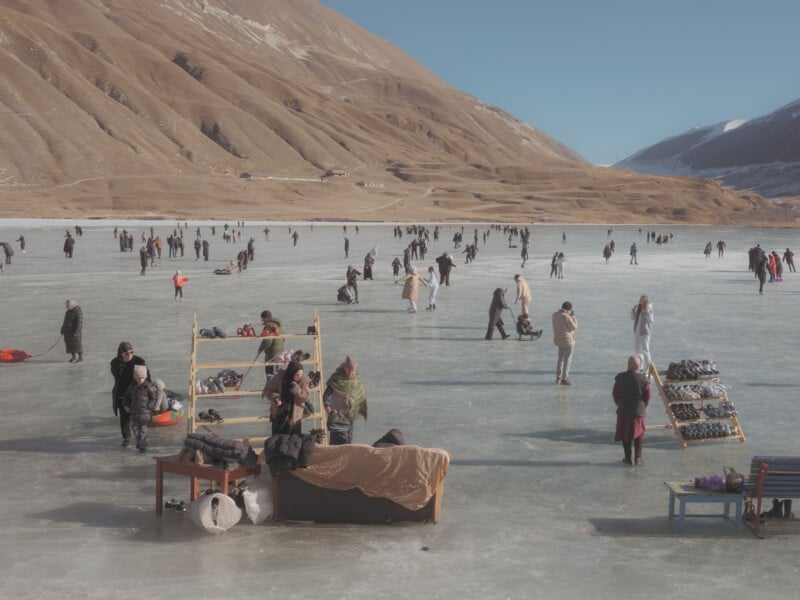 People skate and walk on a frozen lake surrounded by large, barren mountains under a clear blue sky, with rental ice skates displayed on wooden racks.