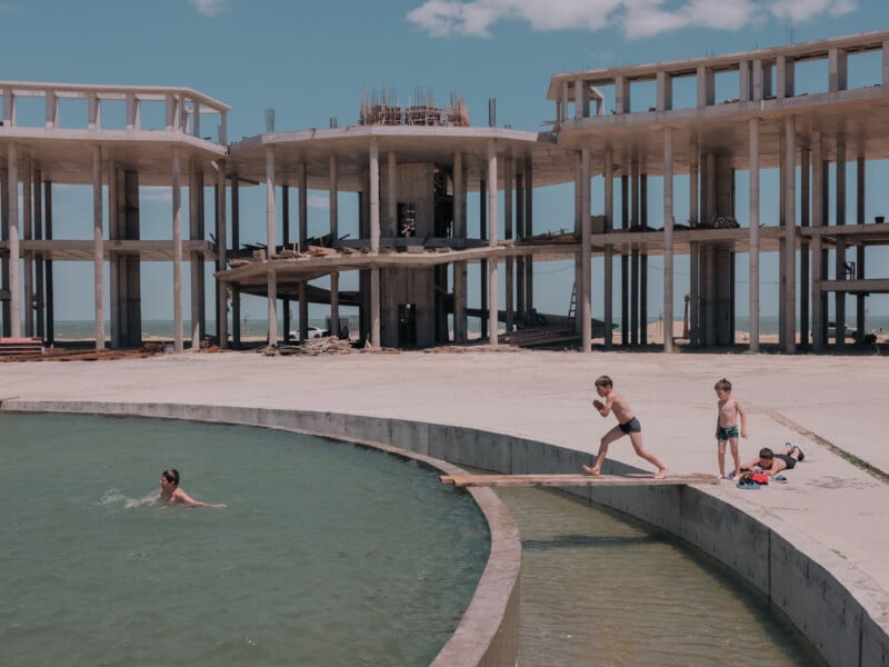Three boys play near a circular pool in front of a large, unfinished concrete building structure.