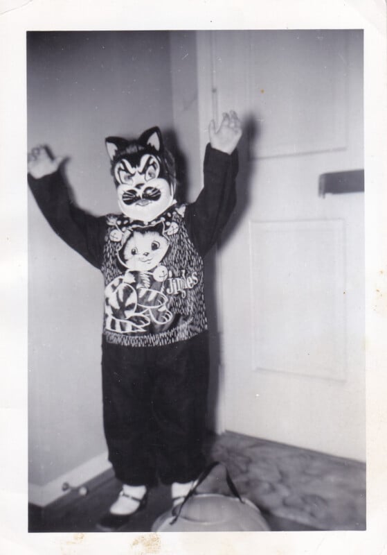 A child in a black cat Halloween costume with a mask and a shirt labeled "Jingles" stands with arms raised, holding a pumpkin bucket.