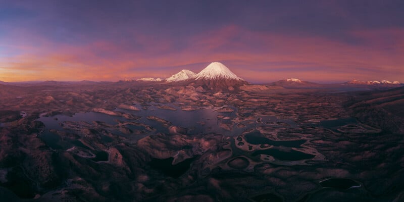 A panoramic view of a snow-capped volcanic mountain rising above a vast, rugged landscape of dark, interconnected lakes at sunset.