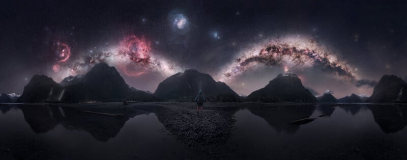 A person stands on a rocky shore between two bodies of water, facing a mountain range under a vibrant, star-filled night sky.