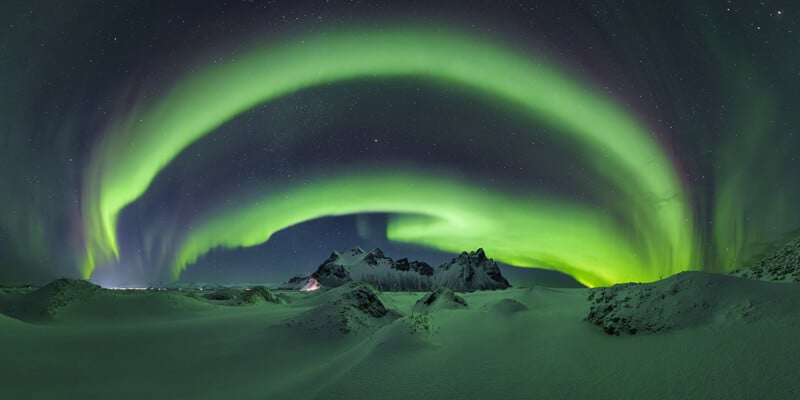 A vibrant green aurora borealis arches across a starry night sky above snow-covered dunes and jagged, dark mountains.