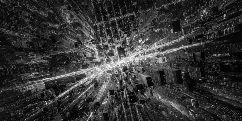 A high-angle, black-and-white aerial view of a dense city at night, featuring a dramatic perspective that converges toward a bright, illuminated street intersection.