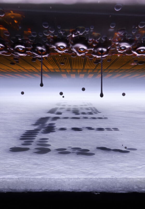 Dark liquid droplets fall from a glowing, grid-patterned surface onto a white, textured plane, creating a trail of dark spots.