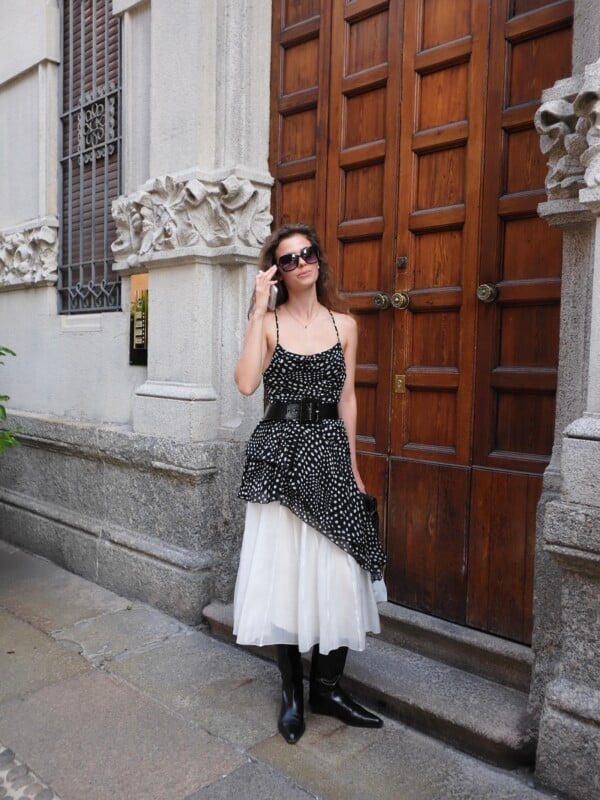 A woman with long brown hair wearing sunglasses, a polka-dot top, a white skirt, and black boots stands in front of a large wooden door while talking on a phone.