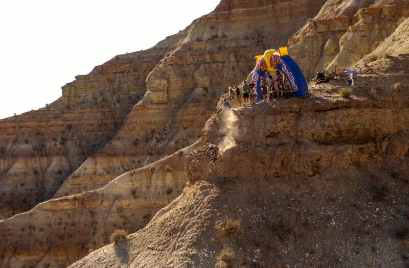 A mountain biker descends a steep, rocky cliff face below a Red Bull branded inflatable arch where a group of spectators is gathered.
