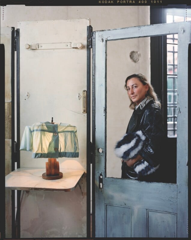 A woman with shoulder-length hair stands behind a weathered, light blue door frame, holding a black and white faux fur accessory.