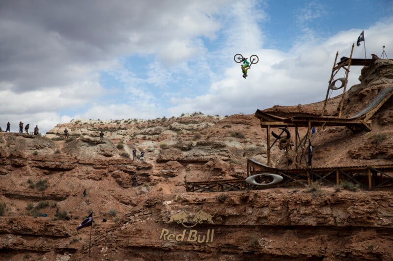 A mountain biker performs a backflip off a wooden ramp in a desert landscape with spectators watching from the surrounding cliffs.