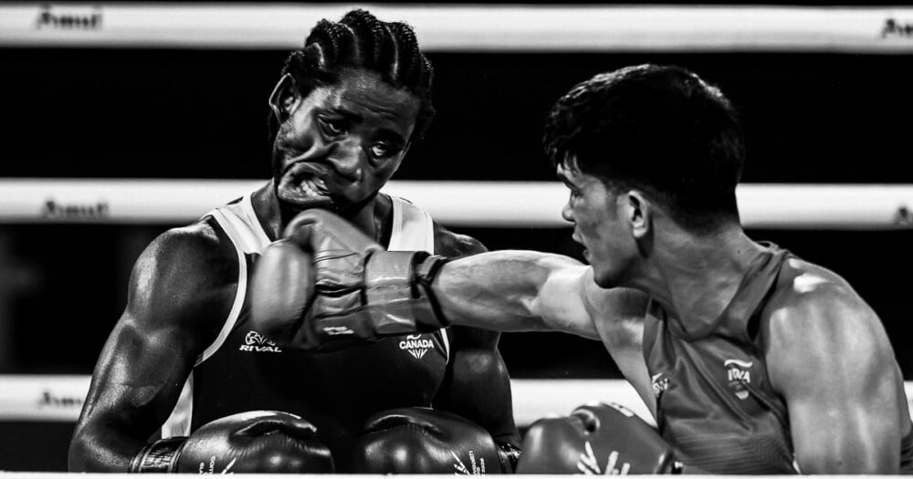 A black-and-white action shot of a boxer landing a punch on his opponent's face during a match.