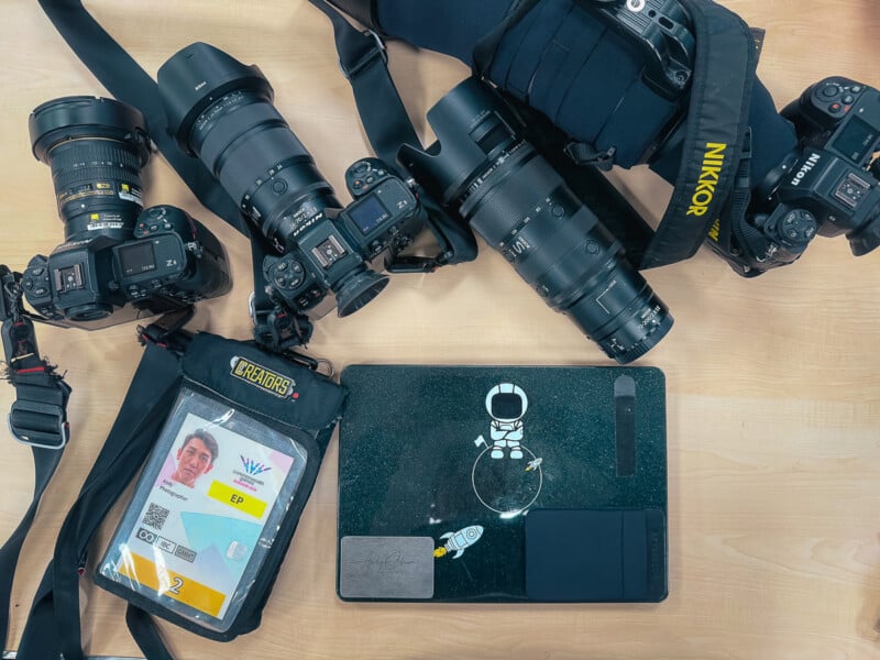 Three Nikon cameras with various lenses, a photographer's credential badge, and a laptop with an astronaut sticker rest on a wooden surface.