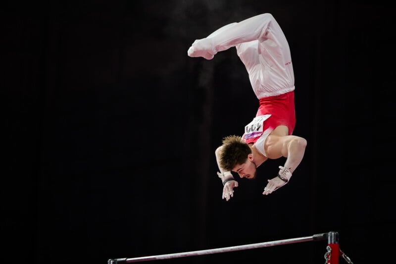 A male gymnast in a red and white uniform performs a release move on the high bar against a dark background.