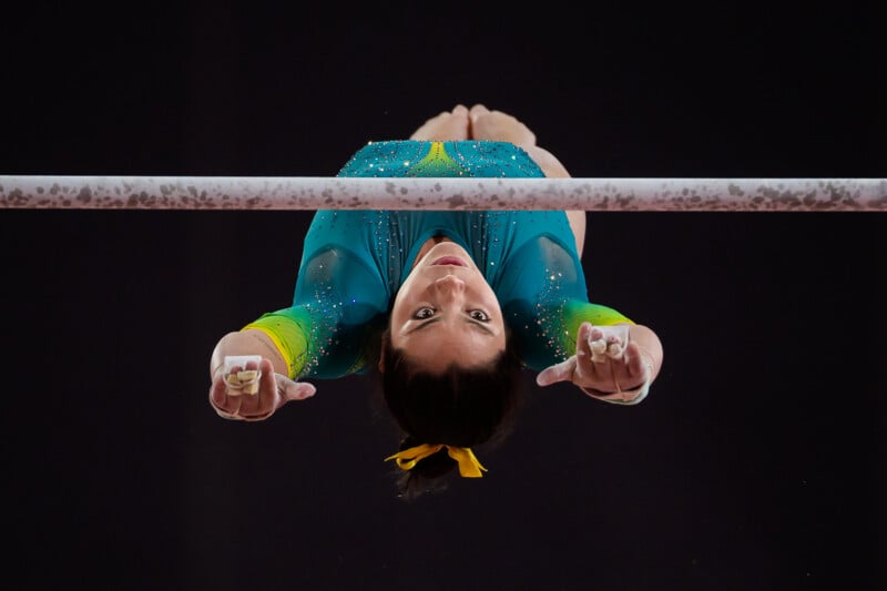 A female gymnast in a teal and yellow leotard performs on the uneven bars, seen from below as she swings over the bar.