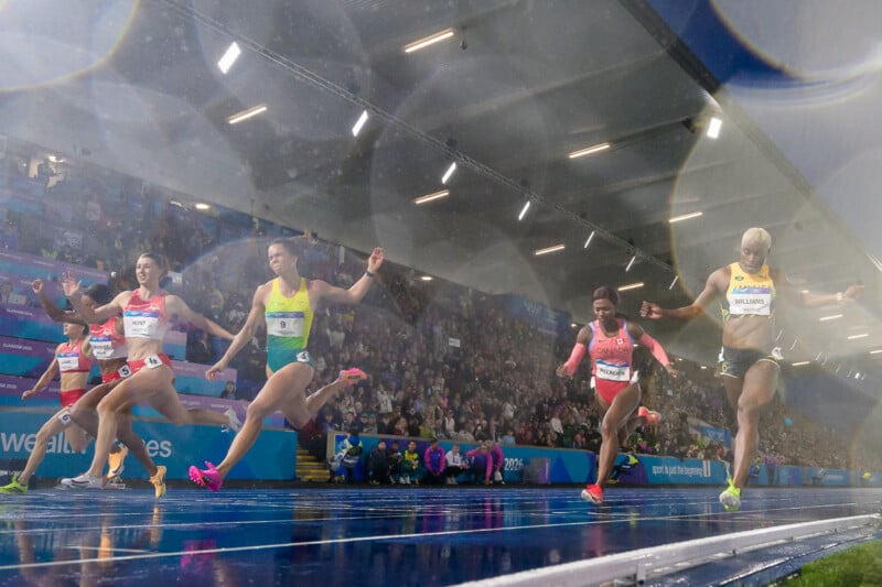 Five female athletes sprint across a wet track toward the finish line in an indoor stadium filled with spectators.