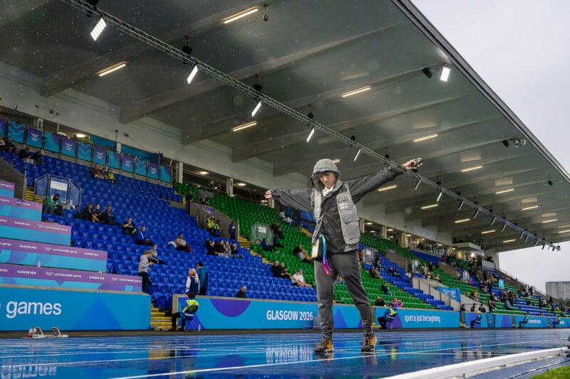 A person in a hooded rain jacket stands on a wet blue running track with arms outstretched, in front of a stadium with blue and green seating.