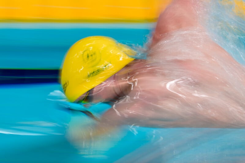 A swimmer wearing a bright yellow cap moves through blue pool water, creating a splash as they perform a freestyle stroke.