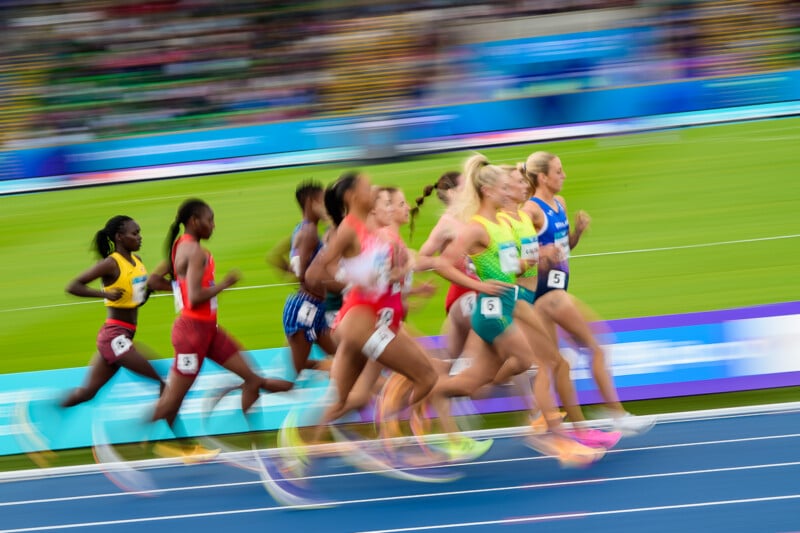 A group of female runners in various colorful uniforms race along a blue track during a track and field event.