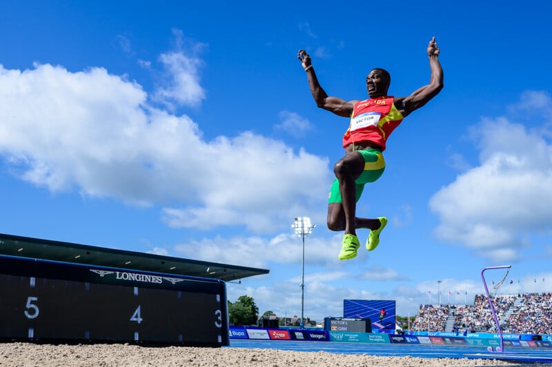 A long jumper from Grenada is mid-air in a jumping pose against a bright blue sky with scattered clouds.