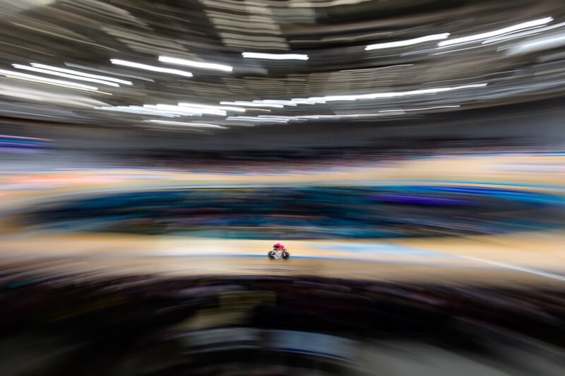 A cyclist in a red jersey rides a track bike on a velodrome, surrounded by a heavy motion blur effect.