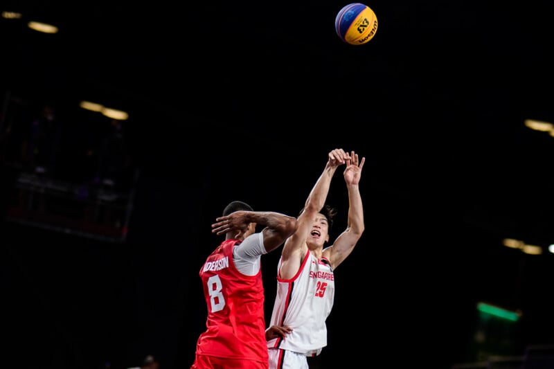 A basketball player in a white Singapore jersey shoots a ball while being defended by a player in a red jersey numbered 8.