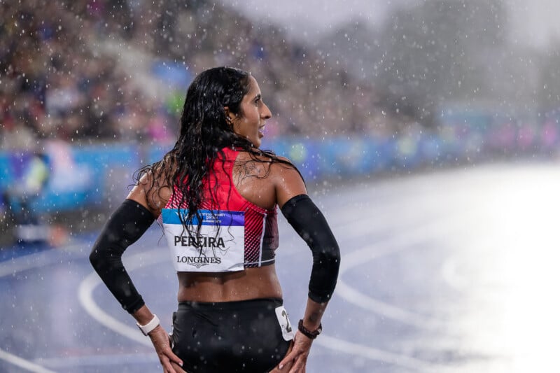 A female athlete with long, wet hair stands on a track in the rain, wearing a red top with a bib labeled "PEREIRA" and black arm sleeves.