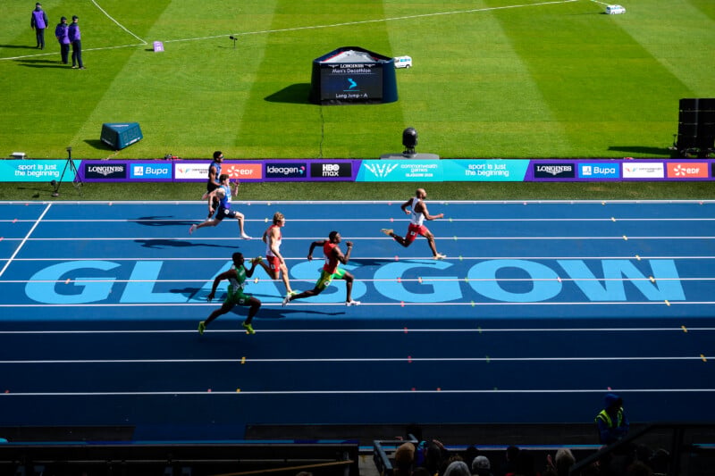 Athletes compete in a track event on a blue track with the word "GLASGOW" printed in large letters, while a digital display shows "Men's Decathlon".
